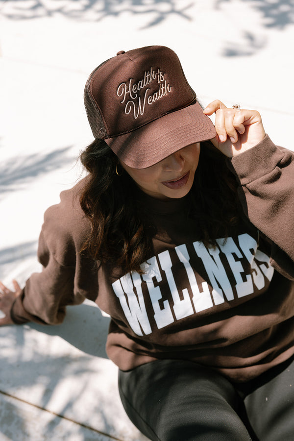 Woman wearing a brown "Health is Wealth Trucker Hat | Lucky Owl" and a matching brown sweatshirt with wellness lettering while sitting outdoors in natural light.