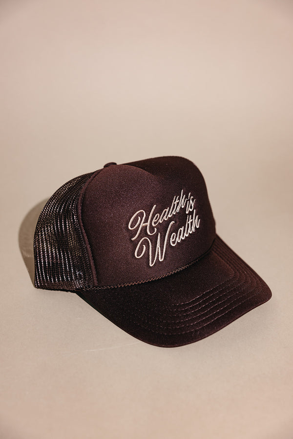 Black mesh-backed trucker hat with white embroidered "Health is Wealth" slogan on the front, styled for casual wellness and outdoor fashion, named Health is Wealth Trucker Hat | Lucky Owl.