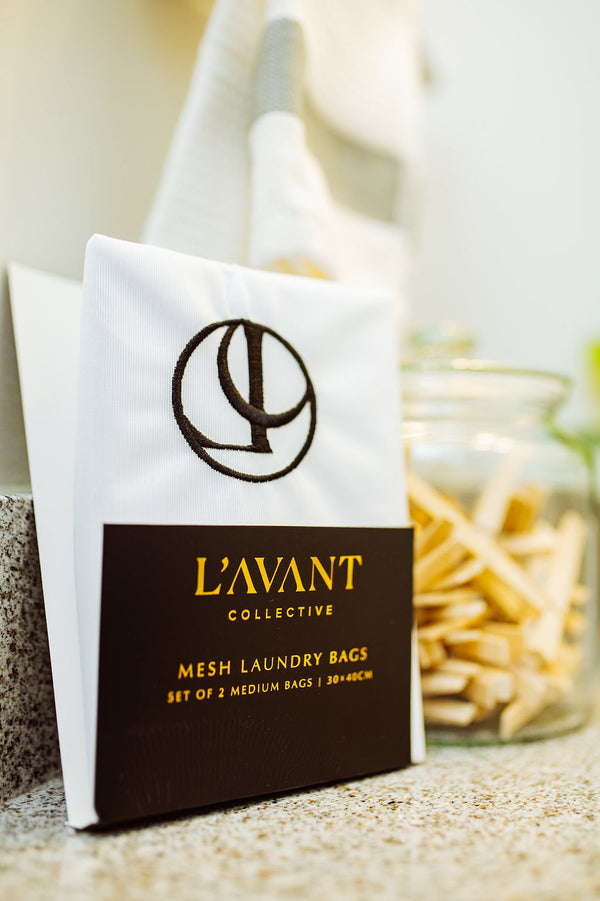 White packaged set of Mesh Laundry Bags Set of 2 | L'AVANT Collective with black and gold branding, displayed on a countertop next to a jar of wooden clothespins.