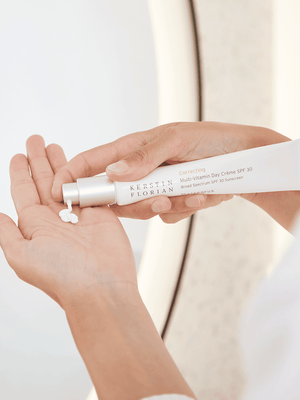 Person dispensing white cream from a tube labeled Correcting Multi-Vitamin Day Creme | Kerstin Florian into their palm, demonstrating skincare application with a neutral background.