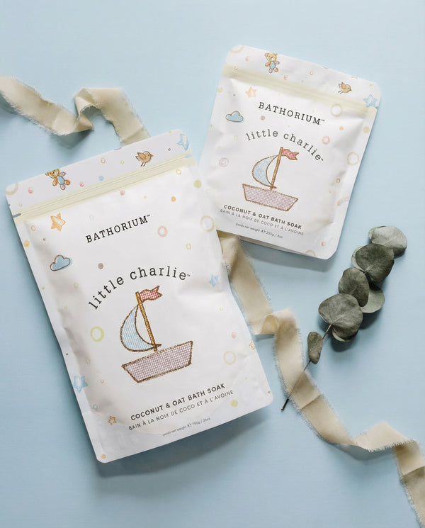 little charlie™ Kids Bath Soak 250g/ 8.8 oz | Bathorium pouches on a blue background with ribbon and smooth stones, showing Coconut and Oat Bath Soak packaging