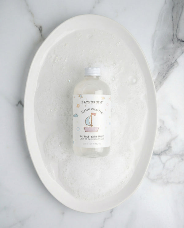 little charlie™ Bubble Bath Milk | Bathorium bottle resting in a white foamy bath in an oval basin on a marble surface