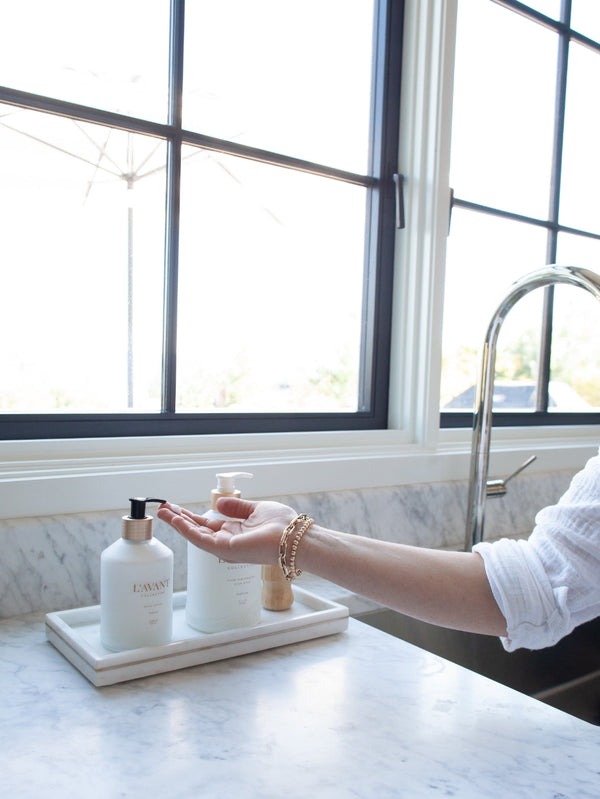 A person dispensing Hand Lotion - Fresh Linen | L'AVANT Collective from a pump bottle on a marble tray next to a kitchen sink in a bright space with large windows, highlighting skincare and body care.