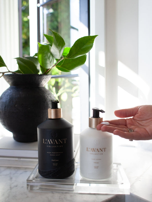 A person dispensing Hand Lotion - Fresh Linen | L'AVANT Collective from a white pump bottle beside a black bottle on a clear acrylic tray, with green leaves and sunlight in the background.