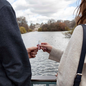 Two people stand by a river, one handing the other a small bottle of Love Therapy Balm | Scentered, suggesting a moment of shared wellness and care outdoors.
