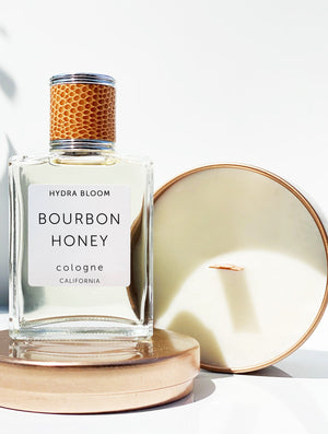 Clear glass bottle of Bourbon + Honey Cologne - Unisex Masculine Scent | Hydra Bloom with honeycomb textured cap, placed on round beige containers in bright natural light