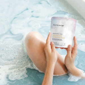 Midnight Superbloom Crush | Bathorium pouch held over a foamy blue bath, with a person relaxing in the water for a soothing body care ritual