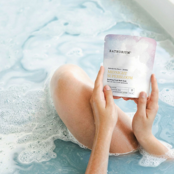Midnight Superbloom Crush | Bathorium pouch held over a foamy blue bath, with a person relaxing in the water for a soothing body care ritual