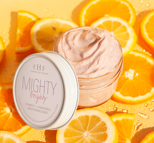 Open jar of Mighty Brighty™ Vitamin C + Chamomile Brightening Mask | Farmhouse Fresh with creamy light pink texture, surrounded by fresh orange and lemon slices on a yellow background.