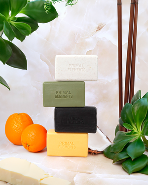 Stacked bars of Luxury African Black Soap | Primal Elements in various natural colors surrounded by fresh green leaves and vibrant oranges, emphasizing skincare and wellness.