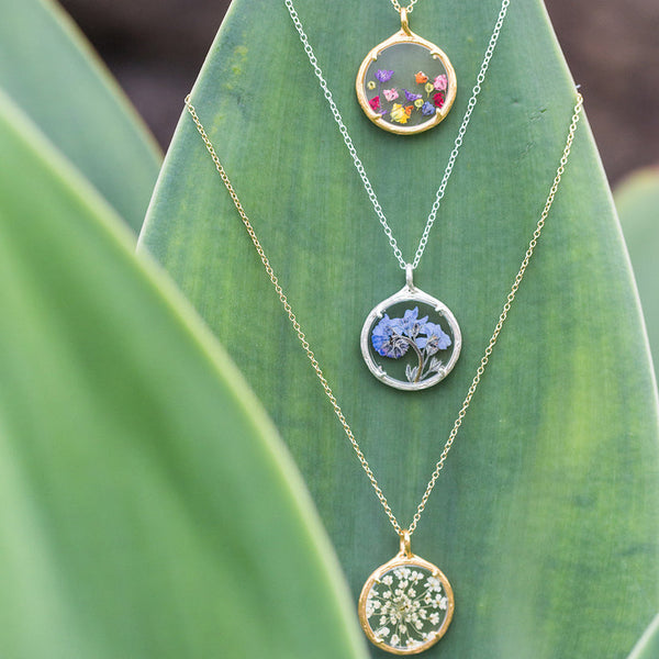 Three Small Botanical Necklace | Catherine Weitzman Jewelry pendants with delicate pressed flowers displayed on a green leaf background.