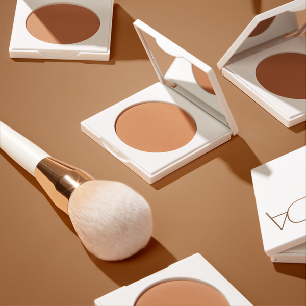 Close-up of Bronzing Powder Brush | NUDA with soft bristles next to open compacts of bronzing powder against a warm tan background, showcasing makeup and skincare essentials.