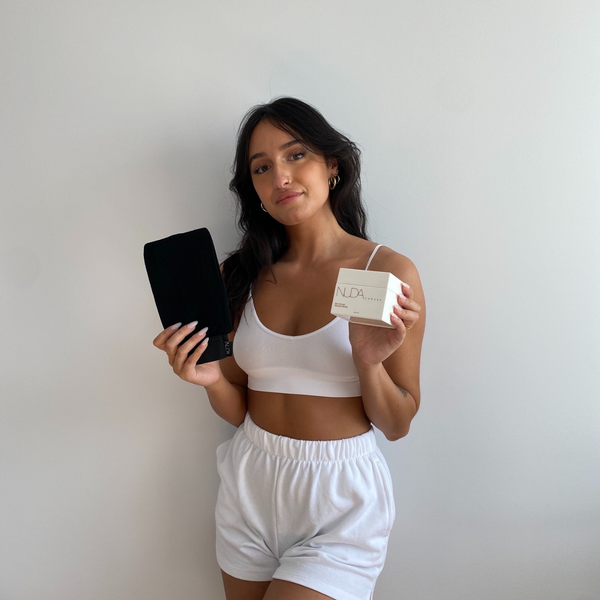 Woman wearing white activewear holding a black exfoliating mitt and a box labeled NUDA, demonstrating skincare and body care wellness products including the Exfoliating Mitt | NUDA.
