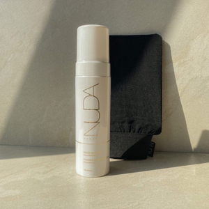 White bottle of skincare product with minimalist NUDA branding standing next to a black exfoliating mitt, highlighting body care and wellness essentials in soft natural light.