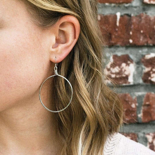 Close-up of a woman wearing silver hoop earrings against a brick wall background, showcasing the Olivia Hoop Earrings | Purpose Jewelry with a minimalist and elegant design.