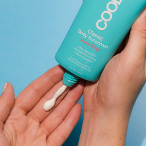 Person applying creamy white lotion from Classic Body Organic Sunscreen Lotion SPF 50 - Guava Mango | COOLA onto hand on a blue background for skincare and body care.