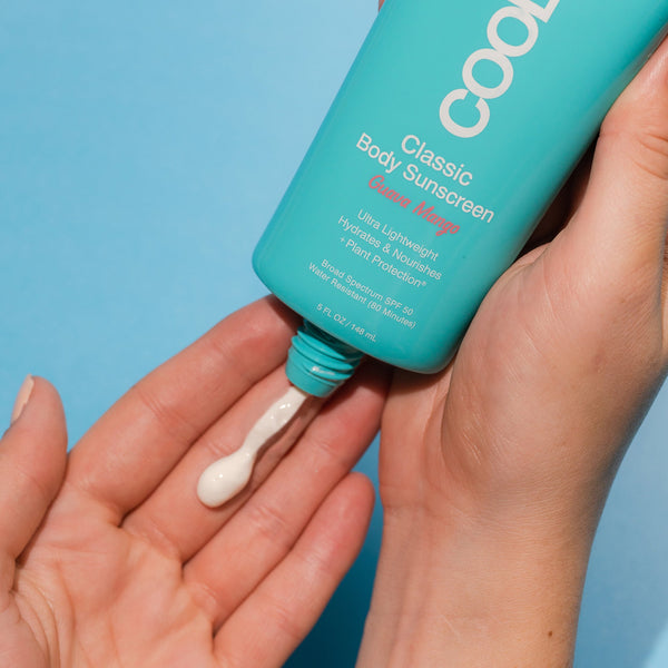 Person applying creamy white lotion from Classic Body Organic Sunscreen Lotion SPF 50 - Guava Mango | COOLA onto hand on a blue background for skincare and body care.