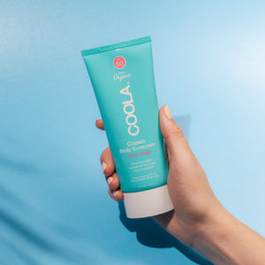 Hand holding a turquoise tube of Classic Body Organic Sunscreen Lotion SPF 50 - Guava Mango | COOLA against a light blue background, showcasing skincare for sun protection and body care.