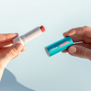Hands holding an open Classic Liplux® Lip Balm Tinted SPF 30 | COOLA with visible tinted balm and turquoise cap against a light background, highlighting skincare and sun protection.