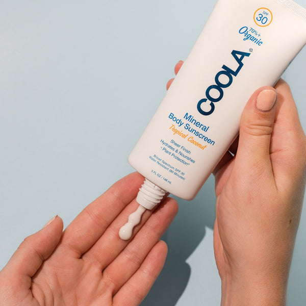 Person squeezing a small amount of Mineral Body Organic Sunscreen Lotion SPF 30 - Tropical Coconut | COOLA onto their hand against a light blue background for body care and skincare wellness.