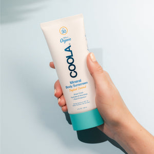 Hand holding a tube of Mineral Body Organic Sunscreen Lotion SPF 30 - Tropical Coconut | COOLA against a light blue background, showcasing skincare and body care product.