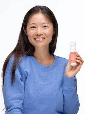 Smiling woman in a blue sweater holding a bottle of Patch Prep Cleanser™ | Dermaclara, promoting skincare and wellness.
