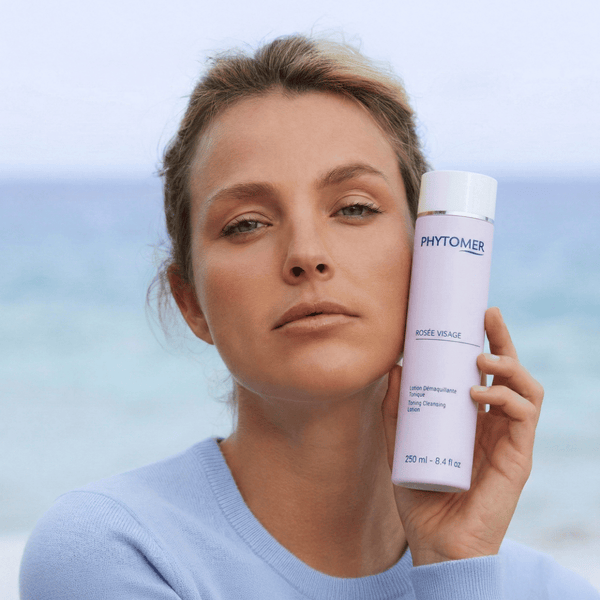 Woman with natural makeup holds a bottle of Rosée Visage (Toning Cleansing Lotion) | Phytomer near her face against a soft ocean background, promoting skincare and wellness.