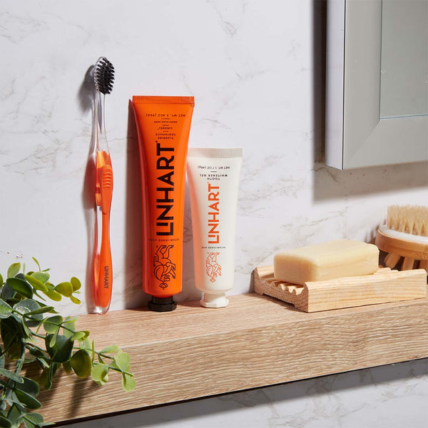 Nano-Silver Toothbrush | LINHART with black bristles and transparent orange handle displayed on a wooden shelf next to LINHART oral care tubes and natural soap in a bright bathroom setting.