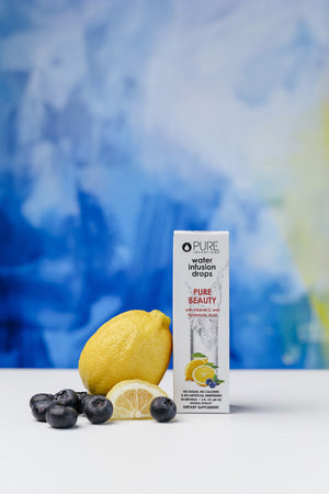 Pure Beauty Water Infusion Drops - Berry Lemonade | Pure Inventions packaging displayed beside fresh lemon and blueberries on a white surface with a soft blue and yellow background, promoting skincare and wellness supplements.