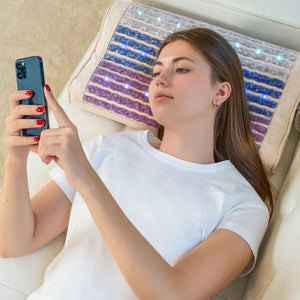 Young woman relaxing on a couch using a smartphone while resting her head on a Rainbow Chakra Pillow Soft | HealthyLine that features calming blue and purple tones with gentle LED lights.