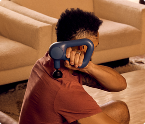 Person using a navy recovery device on their upper back, demonstrating muscle relief with Theragun Relief | Therabody in a cozy living room setting.