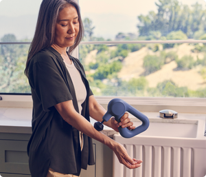 Woman using a navy Theragun Relief | Therabody percussive therapy device on her forearm near a window with a scenic outdoor view, promoting wellness and muscle recovery.