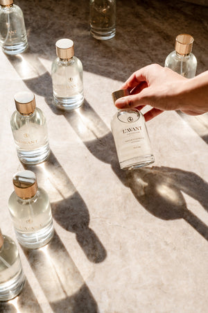 A hand picking up a clear glass bottle with a gold cap of Room Spray | L'AVANT Collective, surrounded by multiple similar bottles casting shadows on a marble surface in natural light.