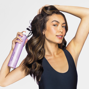 Woman with long wavy hair holding a lavender bottle of Heat Guard Hair Spray | SUTRA, demonstrating hair care and styling with a sleek black tank top.