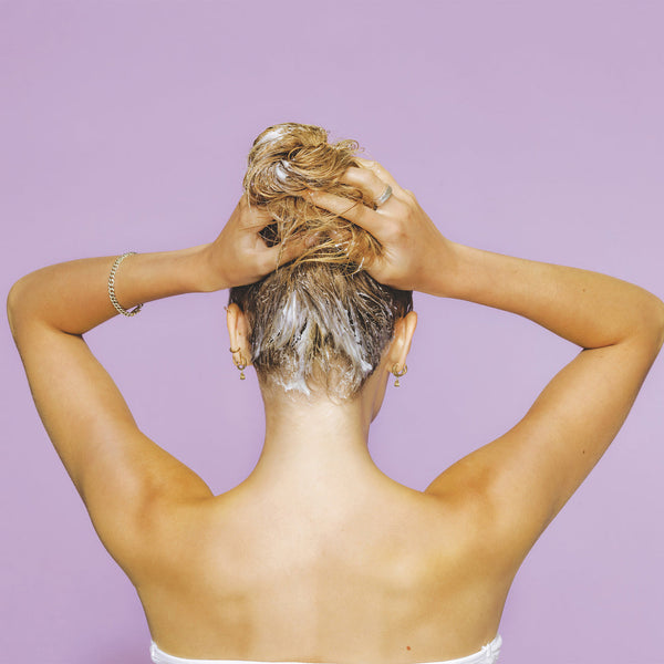 Woman applying Heat Guard Leave In Cream | SUTRA to the roots of her wet hair against a purple background, demonstrating hair care and protection.