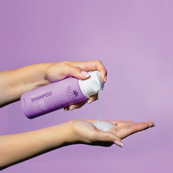 A person dispensing creamy shampoo from a lavender bottle labeled Heat Guard Shampoo | SUTRA onto their palm against a soft purple background.