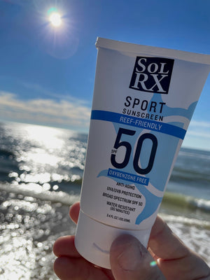 Hand holding a tube of SPORT Sunscreen SPF 50 | SolRX Sunscreen against a bright sunny sky and sparkling ocean background, emphasizing sun protection and reef-friendly skincare.