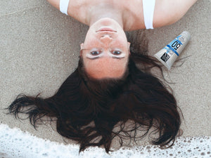 Woman lying on beach sand with wet hair next to a tube of SPORT Sunscreen SPF 50 | SolRX Sunscreen, highlighting skincare protection in a beach setting.
