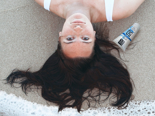 Woman lying on beach sand with wet hair next to a tube of SPORT Sunscreen SPF 50 | SolRX Sunscreen, highlighting skincare protection in a beach setting.