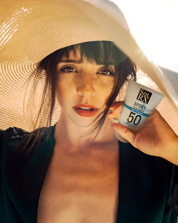 Woman wearing a wide-brimmed sunhat holding a tube of SPORT Sunscreen SPF 50 | SolRX Sunscreen, highlighting skincare and sun protection.