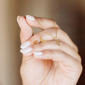 Close-up of a hand holding delicate gold celestial jewelry including star and moon charms, featuring the Galaxy Studs | Purpose Jewelry necklace on a fine chain.