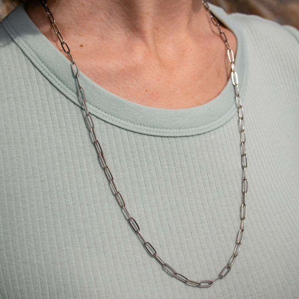 Close-up of a person wearing a Swing Paperclip Necklace - silver tone | Purpose Jewelry, featuring elongated linked chain design against a light green textured top.