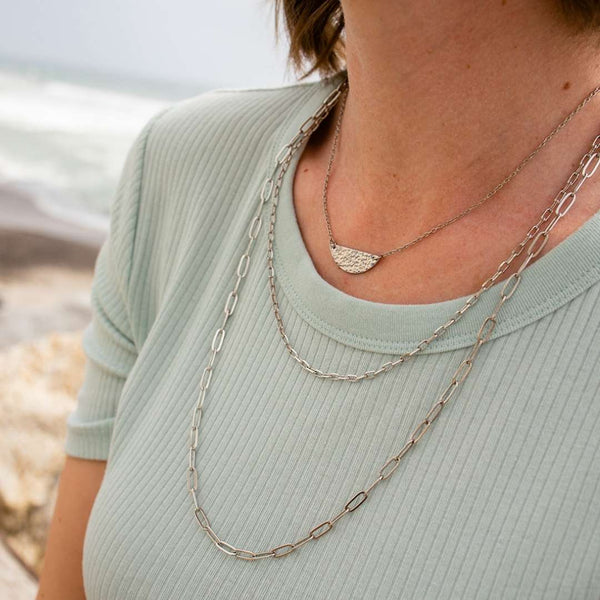 Close-up of a person wearing layered silver necklaces including the Swing Paperclip Necklace - silver tone | Purpose Jewelry over a light green ribbed top near a beach setting.