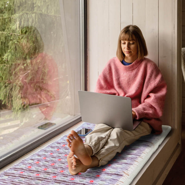 Woman sitting on a window seat using a laptop while resting on the TAJ-Mat™ Full 7224 Firm - Photon PEMF InfraMat Pro® | HealthyLine, showcasing comfort and wellness in a cozy indoor setting.