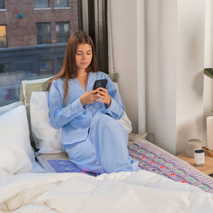 A woman in blue striped pajamas sitting on a bed using her phone while resting on the TAJ-Mat™ King 8076 Firm - Photon PEMF Split Inframat Pro® | HealthyLine wellness device with glowing lights.