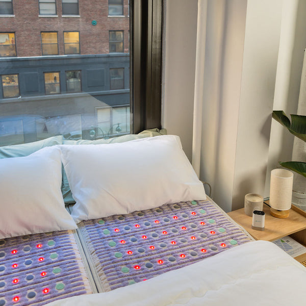 Two white pillows resting on a bed with the TAJ-Mat™ Queen 8060 Firm - Photon PEMF Split Inframat Pro® | HealthyLine placed underneath a white duvet in a bright room with large windows and minimalist decor.