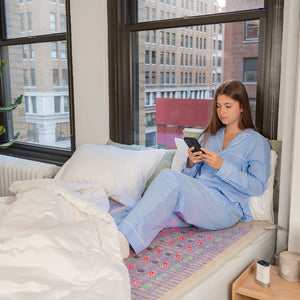Young woman in blue pajamas using a smartphone while sitting on a bed with the TAJ-Mat™ Queen 8060 Firm - Photon PEMF Split Inframat Pro® | HealthyLine for wellness and recovery in a bright urban bedroom.