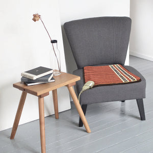A cozy gray chair with a PEMF Far Infrared Heating Mats | HealthyLine placed on the seat, next to a wooden side table holding stacked books and a glass bottle with a dried flower.