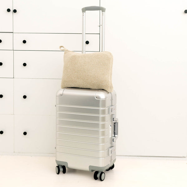 Soft beige blanket from the Travel Set (Blanket, Socks, Eyemask, Bag) | Kashwére draped over a silver carry-on suitcase in a minimalist white room.