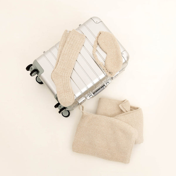 Travel Set (Blanket, Socks, Eyemask, Bag) | Kashwére featuring a cozy beige blanket, matching socks, and eyemask displayed with a silver suitcase, perfect for comfortable travel.